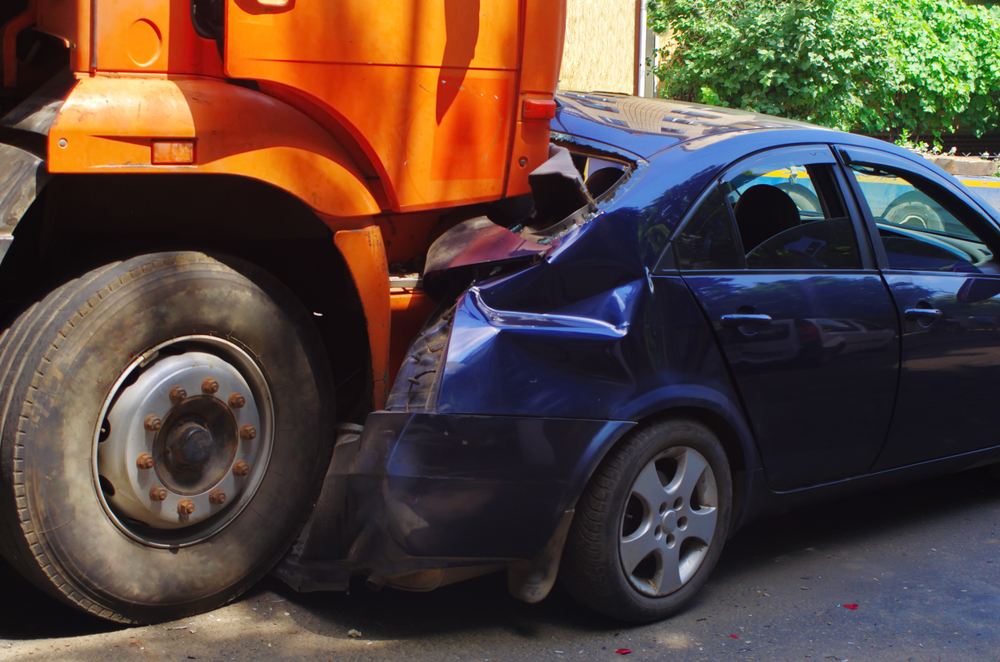 Small blue car getting crushed by large commercial truck in a car accident involving a company vehicle.